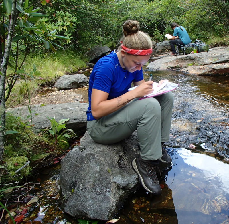 A young woman with a red headband is sitting on a rock by a stream, writing in a notebook. She is wearing a blue shirt, olive-green pants, and hiking boots. The stream is clear and shallow, and there is another person sitting in the background, also writing. The surrounding area is green and lush, with trees and rocks visible.